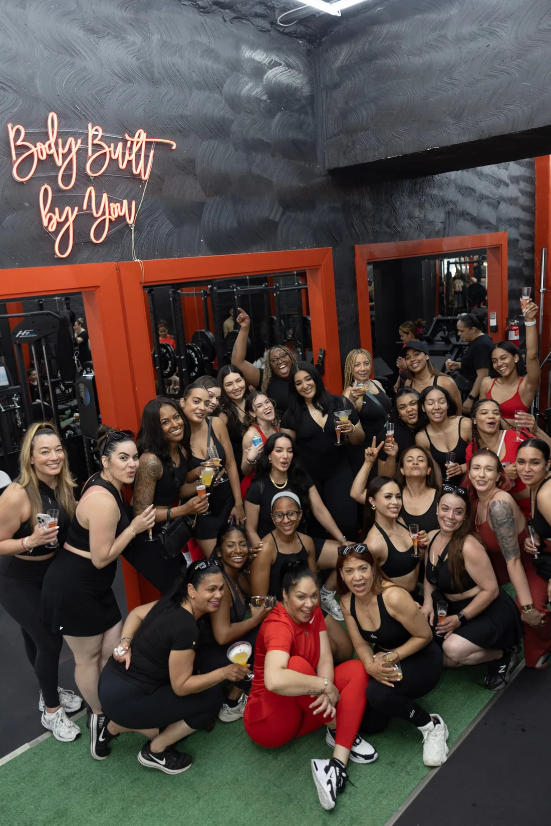 Dozens of women celebrating together inside Body Built By You, under the neon sign and red mirror frames
